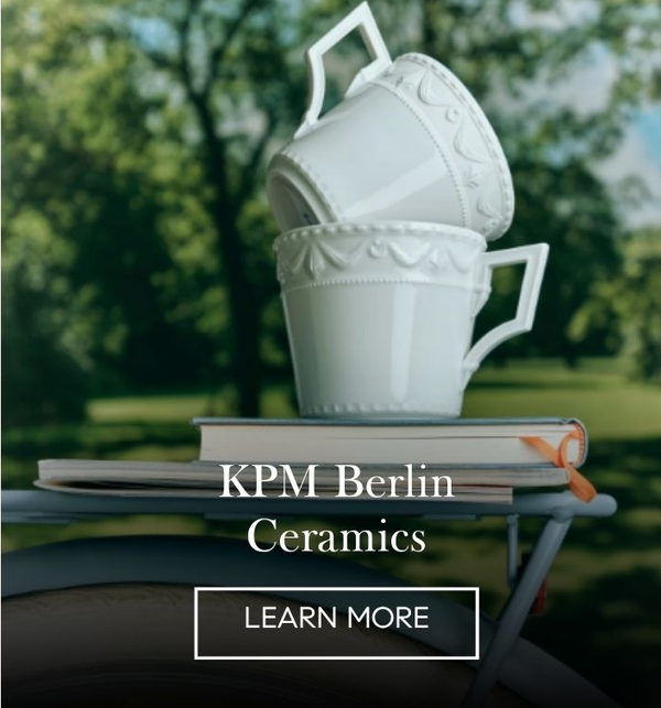 Two white embossed KPM porcelain cups stacked on books on a bicycle rack in a park, 'KPM Berlin Ceramics' with a Learn More button